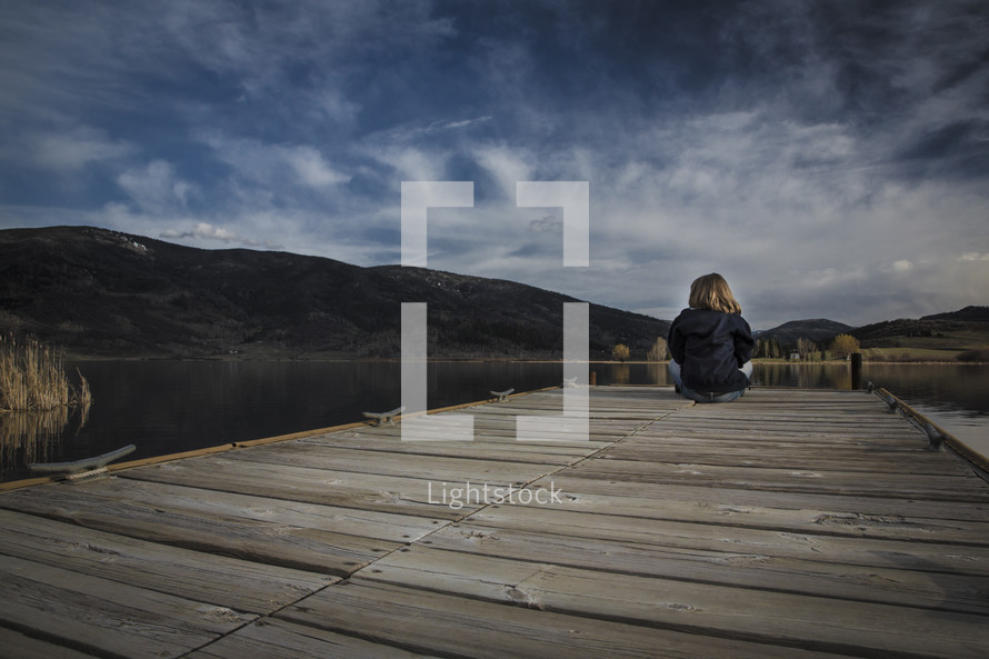A young girl sits at the end of a pier facing clouds and mountains.