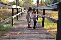 brother and sister holding hands outdoors on a wooden foot bridge 
