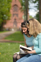 Young women smiling while reading in the bible together outside on a sunny day with a cathedral in the background. 