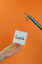 a hand grabbing for a piece of paper with the word LUCK on it hanging from a fishing line 