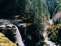 A waterfall on a mountain side, trees and a stream in the distance