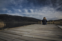 A young girl sits at the end of a pier facing clouds and mountains.