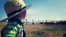Boy in straw hat looking at pasture with herd of cows in the distance.