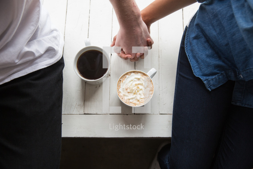 A man and woman sitting and holding hands by two cups of coffee.