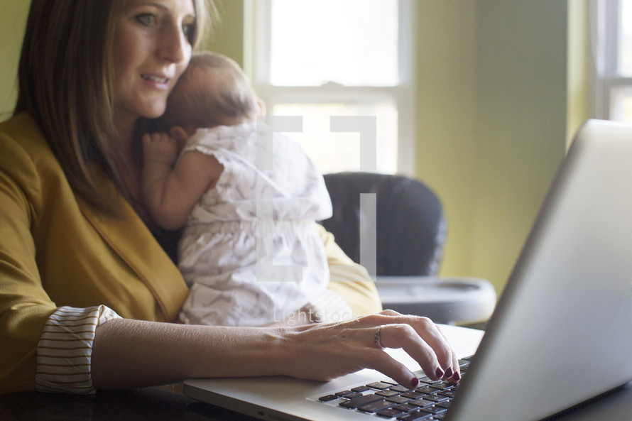 working mom, mother typing on a computer while holding her infant 