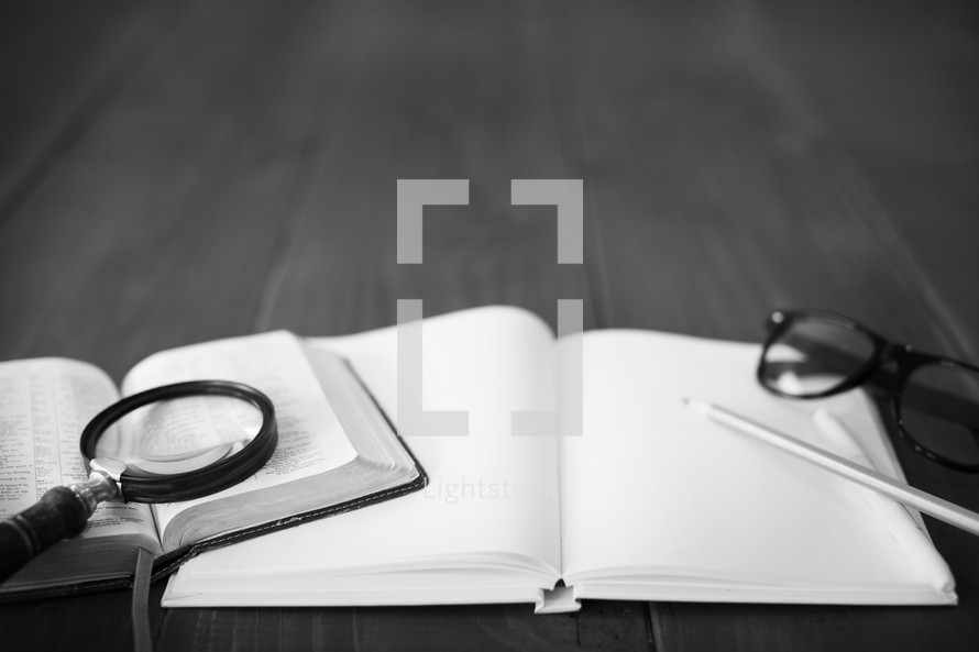 magnifying glass on the pages of a Bible and a journal and pencil and reading glasses 