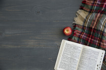 apple, plaid blanket, and open Bible on gray wood boards 