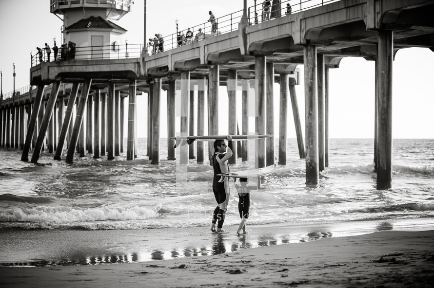 father and son carrying surfboards under a pier 