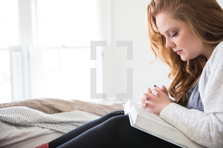 young woman sitting in bed praying over a Bible