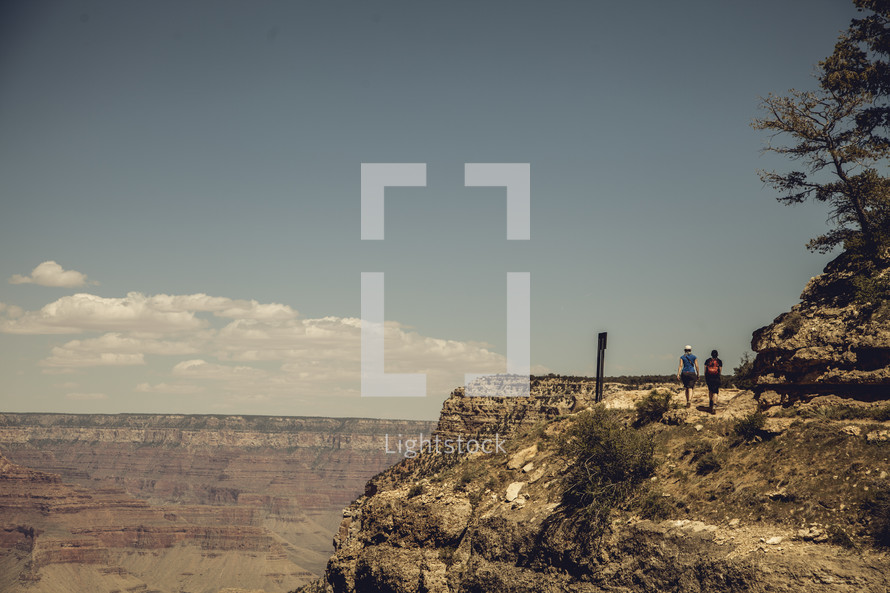 Two people walking along path on a rocky cliff overlooking a canyon.