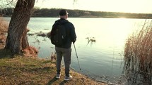 Senior man stand with trekking poles in their hands on the lake, resting after Nordic walking, enjoy nature at sunset time.