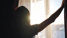 Beautiful woman opening curtains and looking through the window. Sunrise or sunset behind the window. Young woman opening curtains in a bedroom. Positivity and energy concept.