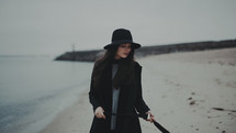 a young woman in a trench coat and hat standing on a beach shore 
