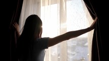 Woman opening curtains and looking through the window. Sunrise or sunset behind the window. Young woman opening curtains in a bedroom. Positivity and energy concept.
