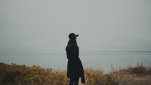 a young woman in a trench coat and hat standing on a coastal shore 