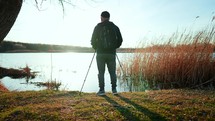 Rear view senior man stand with trekking poles in their hands on the lake, resting after Nordic walking, enjoy nature at sunset time.