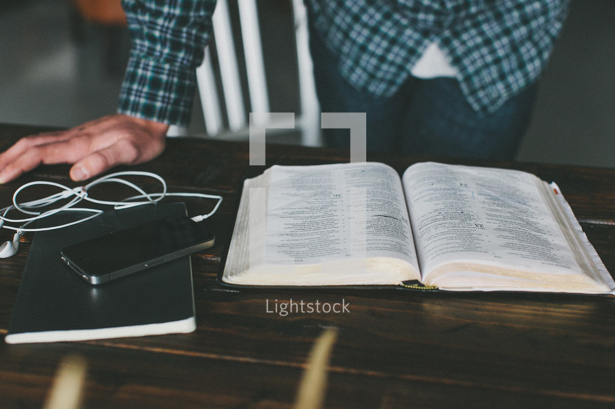 Man's hand leaning on wooden desk with iPod reading open Bible.