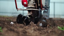 Home farming. Close-up farmer plows the land with a gasoline plow in the garden. Agriculturist controlling two wheel tractor plowing on soil field in plantation. Manual motor plow throwing soil.