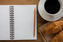 open notebook, pencil, coffee cup, and croissants 