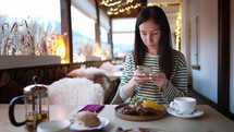 Pretty woman sitting on the cafe terrace on the background of winter mountain landscape, using a smartphone. Woman tourist uses internet on smartphone on restaurant in mountain. Amazing mountains view.