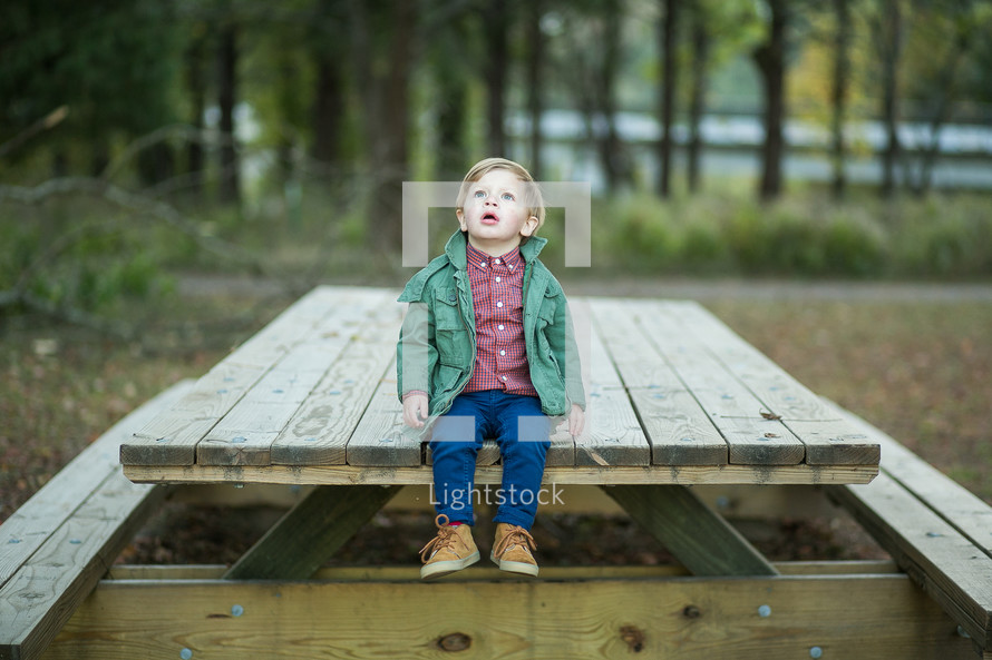 a toddler boy outdoors in a jacket sitting on a picnic table 