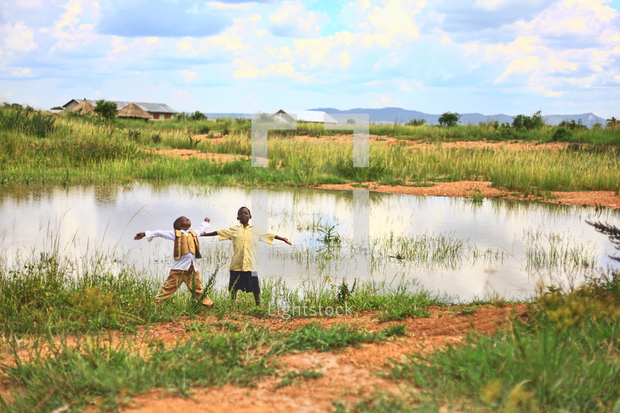 Boys with arms spread standing on the edge of a pond by a village, looking toward the sky.