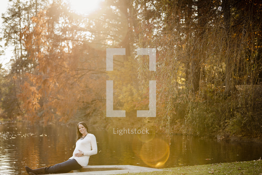 pregnant woman sitting in front of a lake with her hands on her pregnant belly