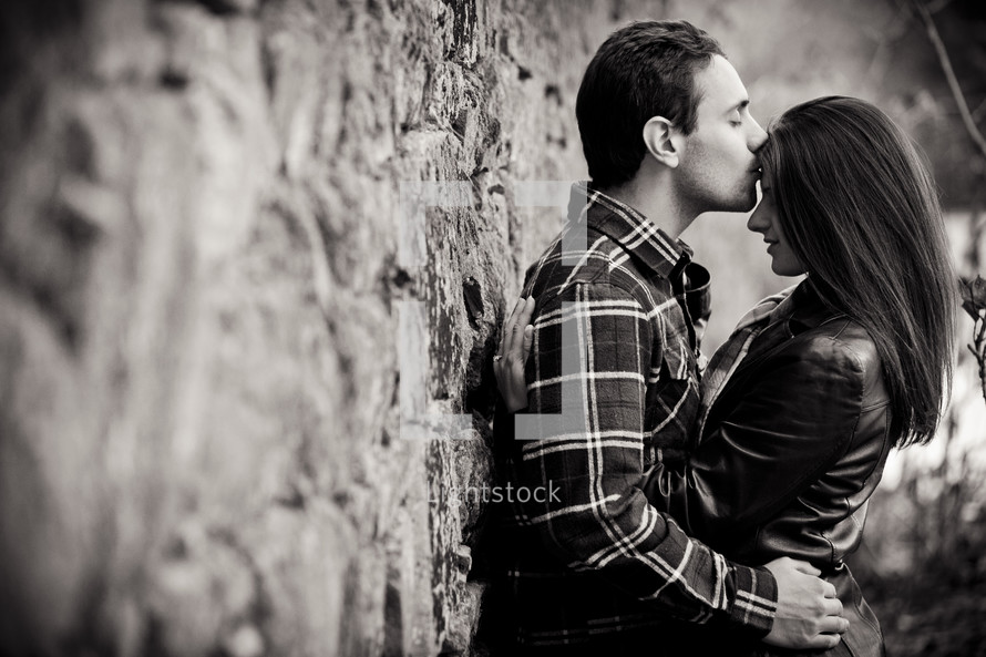 Man and woman leaning against stone wall, embracing with a kiss.