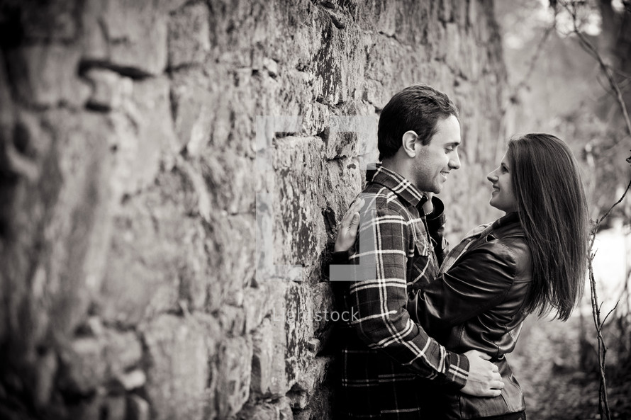 Man and woman leaning against stone wall smiling and embracing.