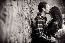 Man and woman leaning against stone wall, embracing with a kiss.