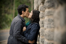 man kissing a woman leaning against a stone brick wall