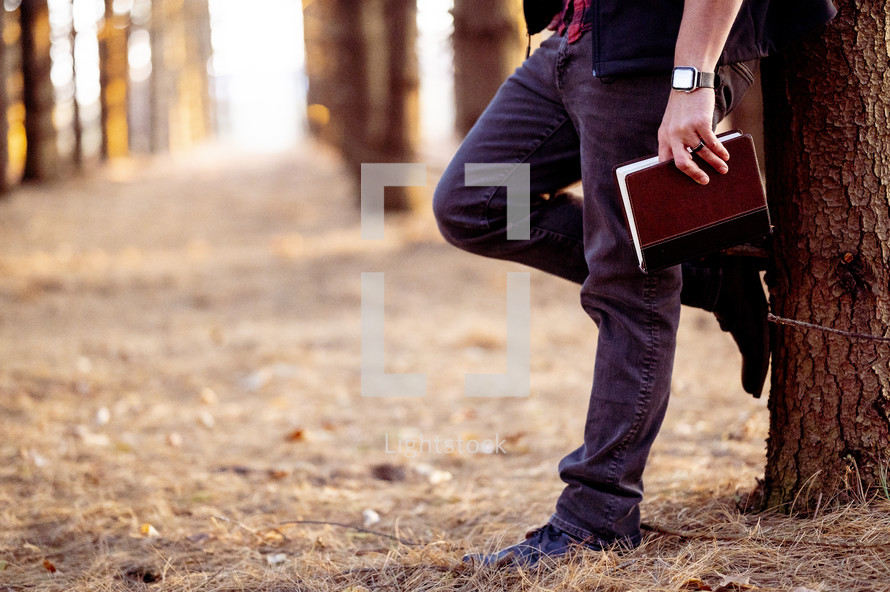 a man in a forest leaning against a tree and holding a Bible in fall 