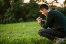a man squatting outdoors with praying hands 