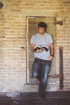man leaning against a wall reading a Bible 