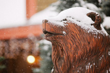 Wood carving of bear head in snow with lighted lodge in the background.