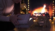 Young woman opening bright gift box near Christmas tree in front of fireplace. Christmas giving concept.
