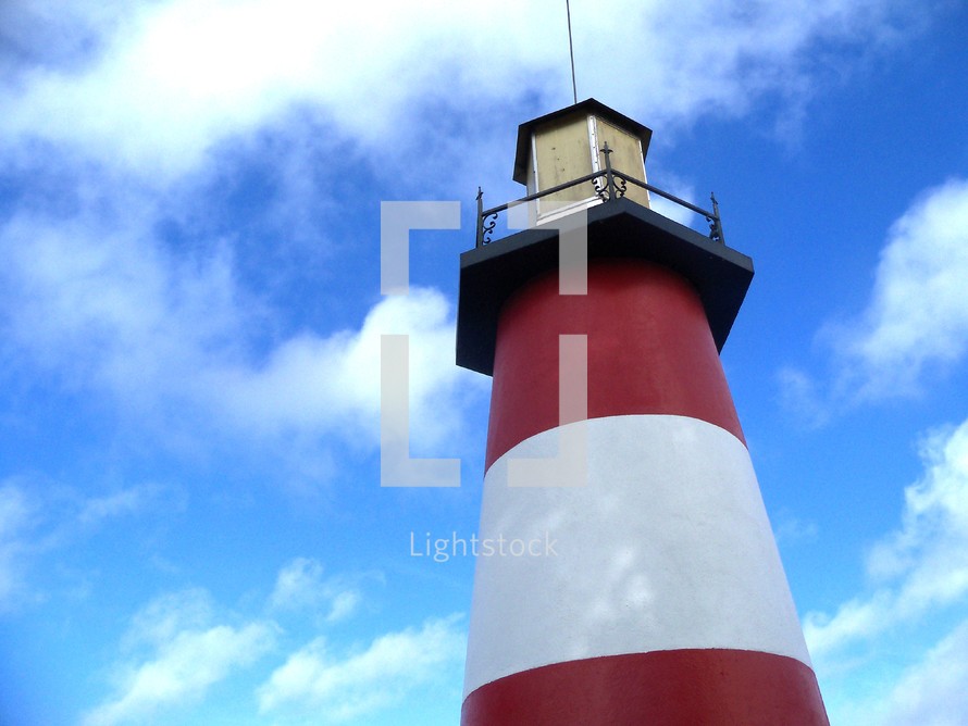 A red and white striped lighthouse against a bright blue sunny sky looking out over a body of water providing safe harbor for incoming boats, ships and water craft. 