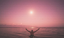 a man with raised arms standing in the ocean at sunset 