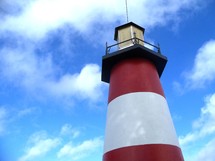 A red and white striped lighthouse against a bright blue sunny sky looking out over a body of water providing safe harbor for incoming boats, ships and water craft. 