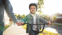 Boy cycling in park with mother who is teaching him to ride bicycle. Young boy is riding for the first time on a sunny street. Happy young family, motherhood and childhood, active lifestyle concept.