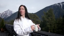 Young woman in a white coat, drinking morning coffee on terrace of hotel, looking at beautiful winter mountains. Woman drinking her morning coffee and enjoying view of mountains. Vacation lifestyle.