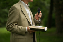 A man holding an open Bible while speaking into a microphone.