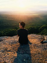 man sitting on a mountaintop at sunrise looking out 