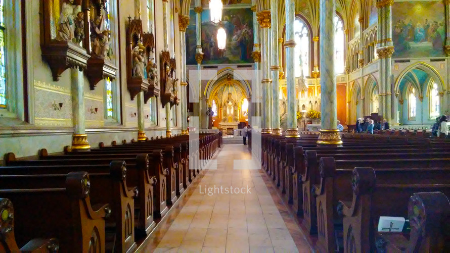 A historic church view of the center aisle and pews lined on each side of the aisle and walls adorned with stained glass windows, sculptures and three dimensional artwork dating back from the early 1800s. 