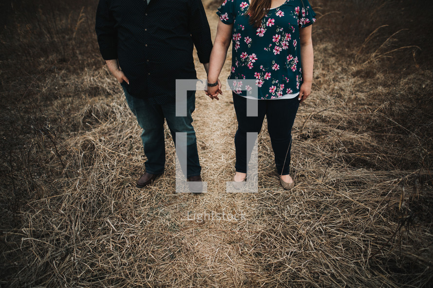 a couple standing in a field of dry grass holding hands 