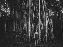 A woman stands in front of an enormous tree in the forest.