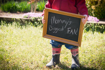 toddler boy holding a chalkboard sign that reads mommy's biggest fan 