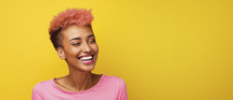 Joyful African American woman with vibrant pink hair laughing against a yellow background.