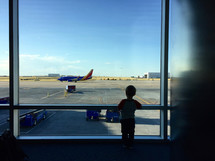 a boy looking out a window in an airport at airplanes on a runway 