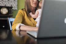 working mom, mother holding her infant daughter while typing on a laptop 